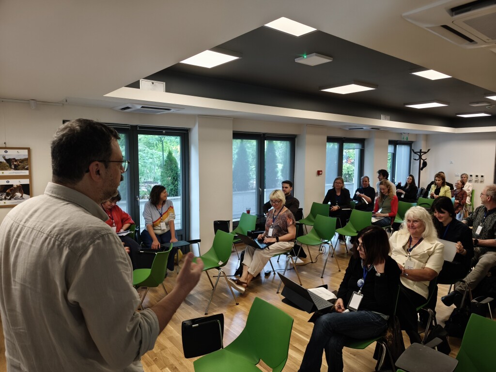 A person is presenting to a group of around 20 participants in a bright seminar room. The audience is seated on green chairs, with some using laptops or taking notes. Large windows overlook a green outdoor area.