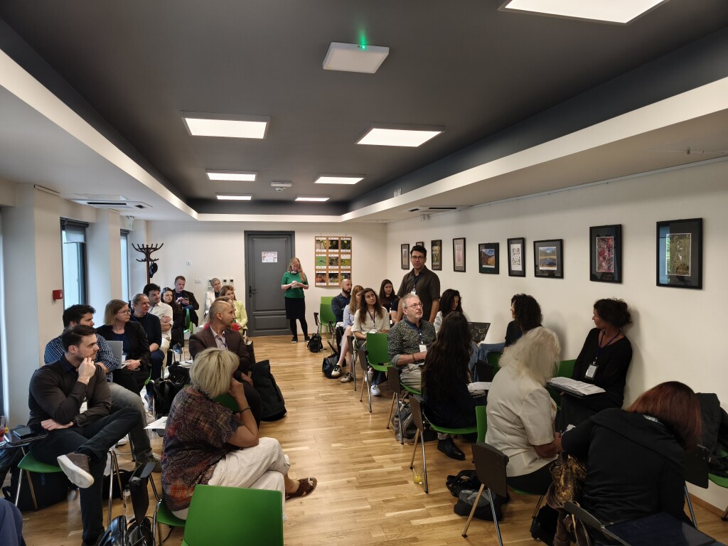 Workshop participants sit in a rectangular chair arrangement and engage in a group discussion. One participant stands and speaks while the others listen attentively. Framed pictures are displayed on the walls.