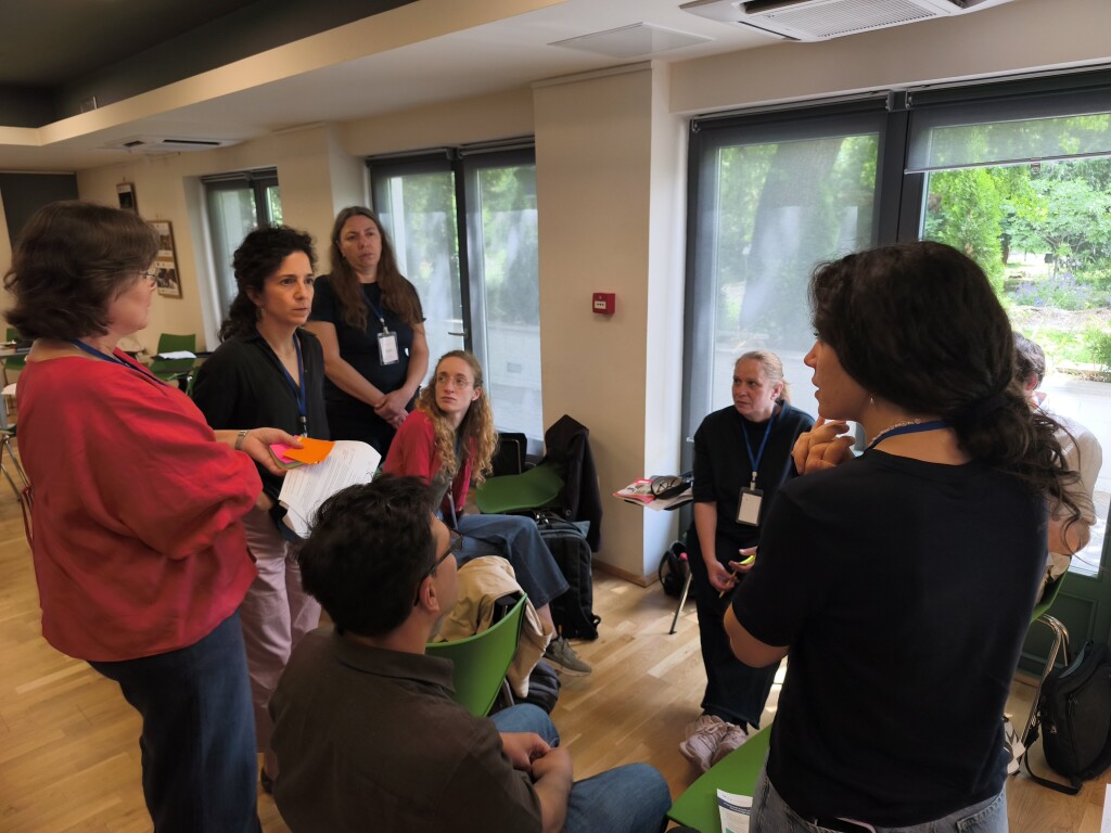 A small group of workshop participants is engaged in discussion near large windows in a well-lit room. One participant holds notes and colorful cards while others listen and contribute to the conversation.