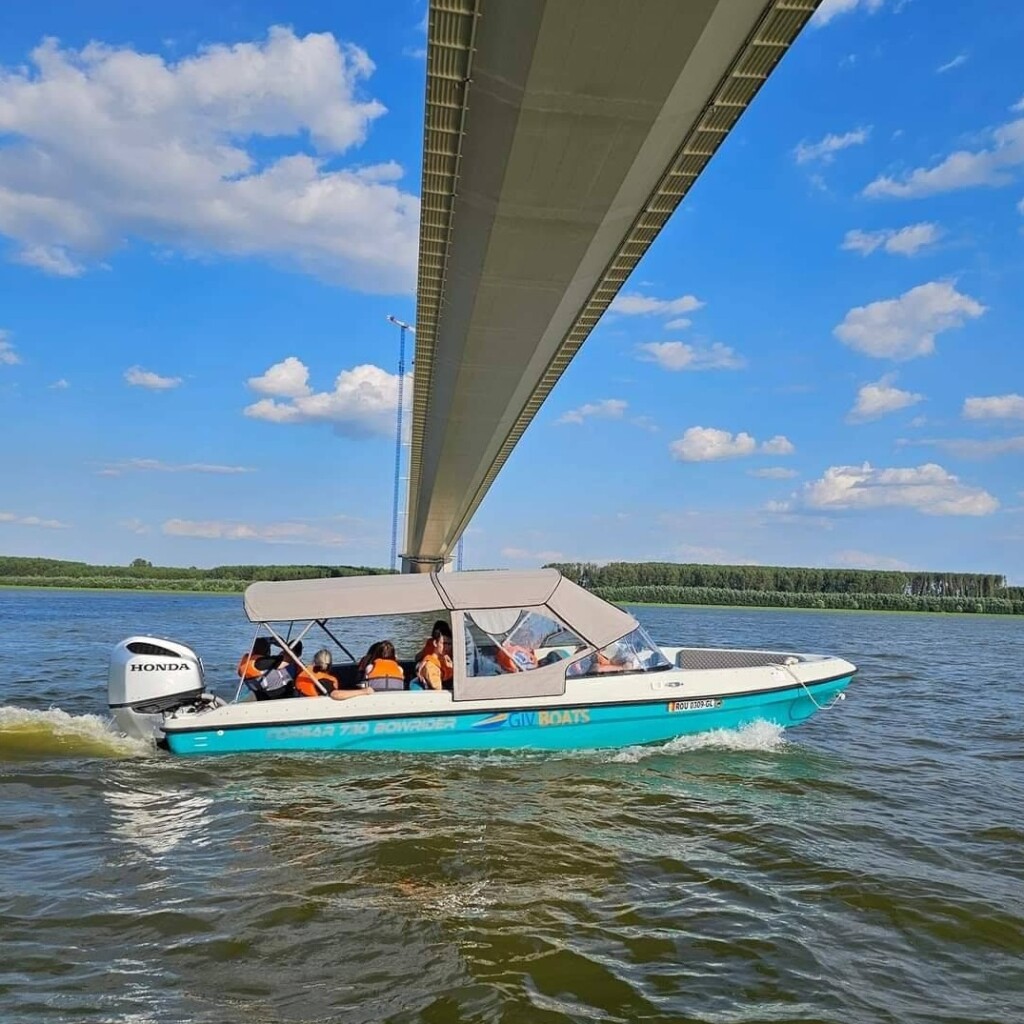 Motorboat travelling on the Danube beneath a large bridge on a clear summer day.