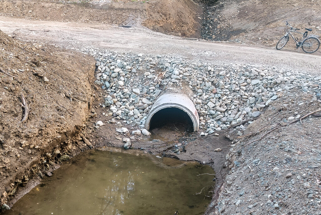 Concrete culvert beneath a gravel road with a small stream flowing through the pipe.