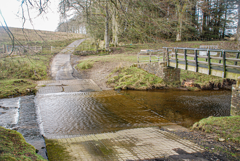 Shallow stream crossing at a paved ford next to a small pedestrian bridge in a rural landscape.