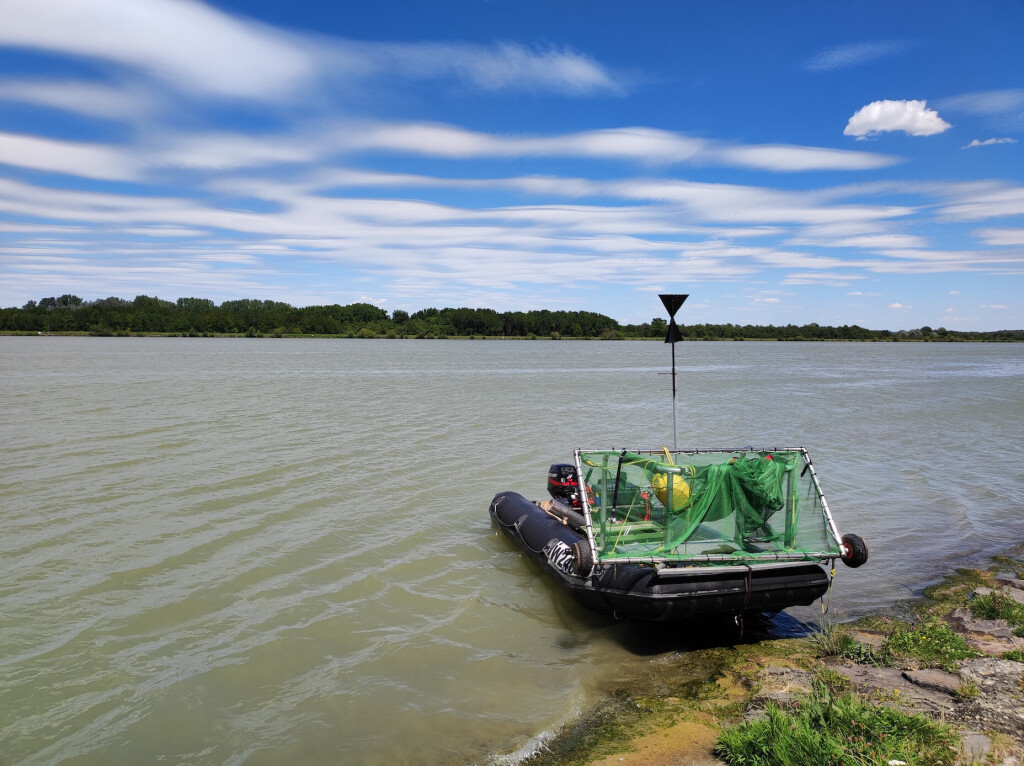 Inflatable boat with research equipment resting on the bank of a river in a natural setting.