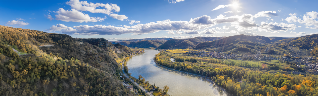 Landscape of a river in between hills with trees and a little village on a sunny day.